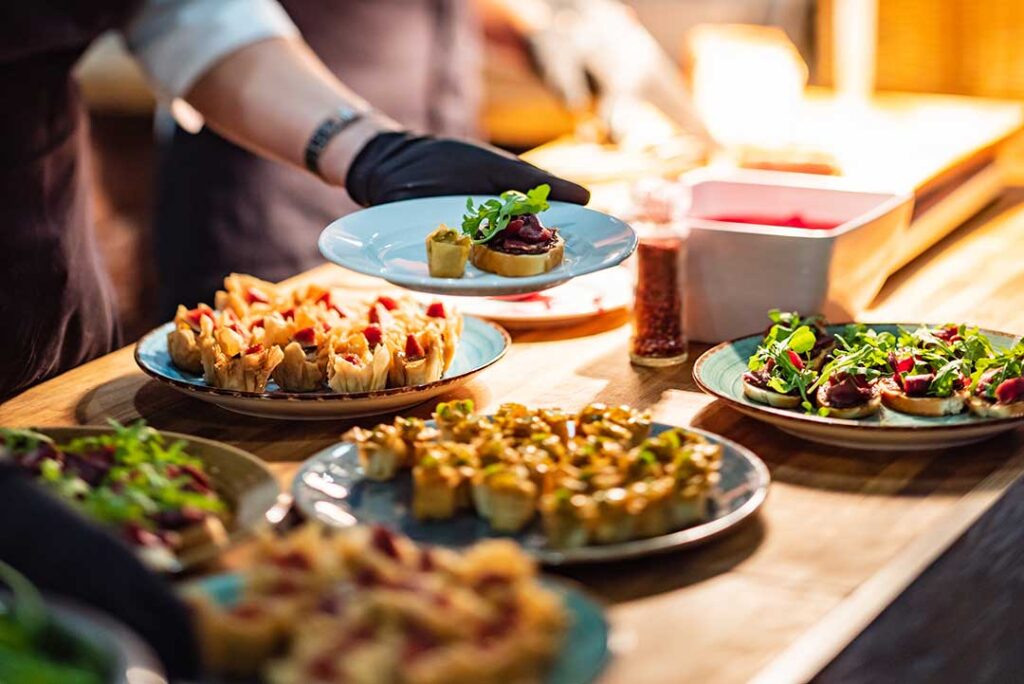Guests serve themselves from a buffet table with covered dishes, plates, drinks, and a printed event budget nearby
